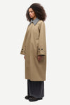 A model wearing a mid-length beige trench coat with a contrasting gray collar, paired with dark wide-leg trousers and black shoes, shown from a side profile against a light gray background.