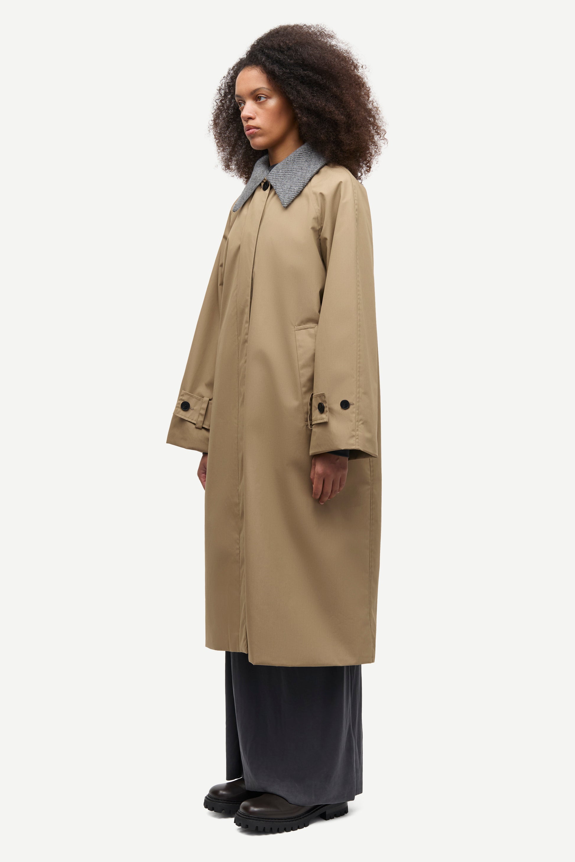 A model wearing a mid-length beige trench coat with a contrasting gray collar, paired with dark wide-leg trousers and black shoes, shown from a side profile against a light gray background.