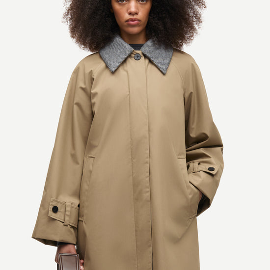 A model with dark curly hair wearing a long beige trench coat with a contrasting gray collar, holding a brown leather bag against a light gray background.