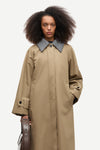A model with dark curly hair wearing a long beige trench coat with a contrasting gray collar, holding a brown leather bag against a light gray background.