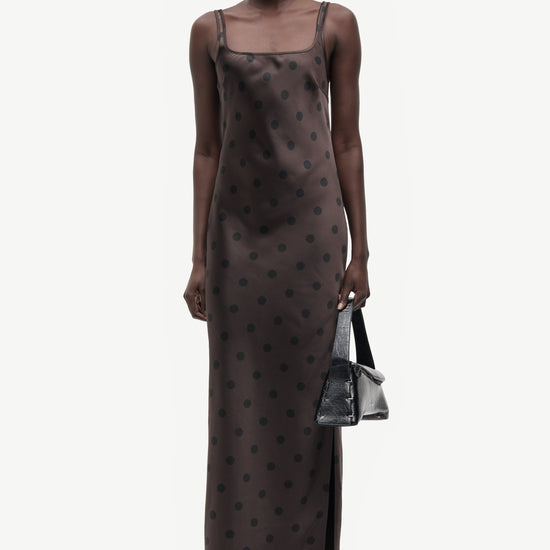 A model wearing a dark brown sheer mesh slip dress with a black polka dot pattern, holding a small black handbag against a light gray background.