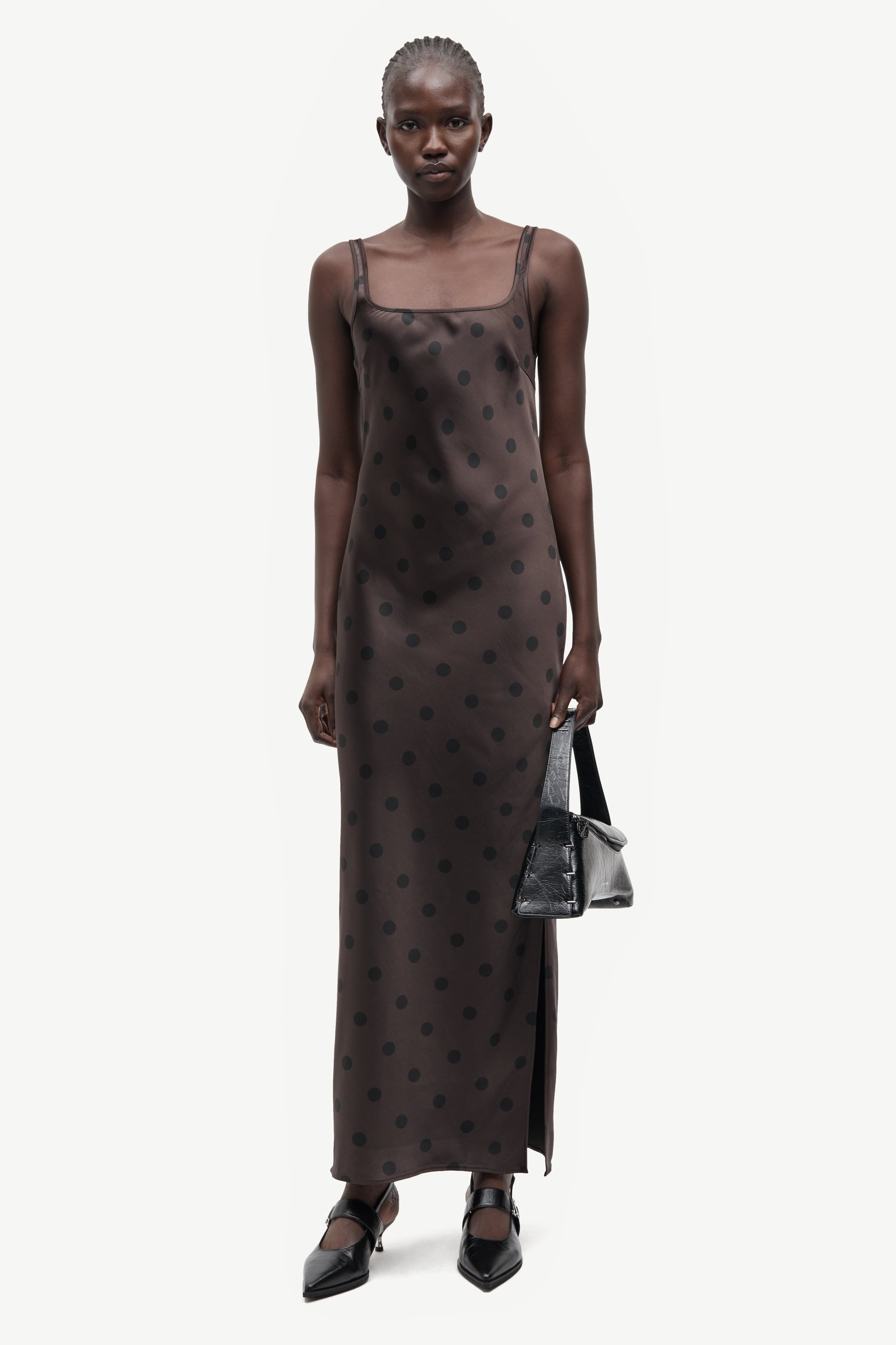 A model wearing a dark brown sheer mesh slip dress with a black polka dot pattern, holding a small black handbag against a light gray background.