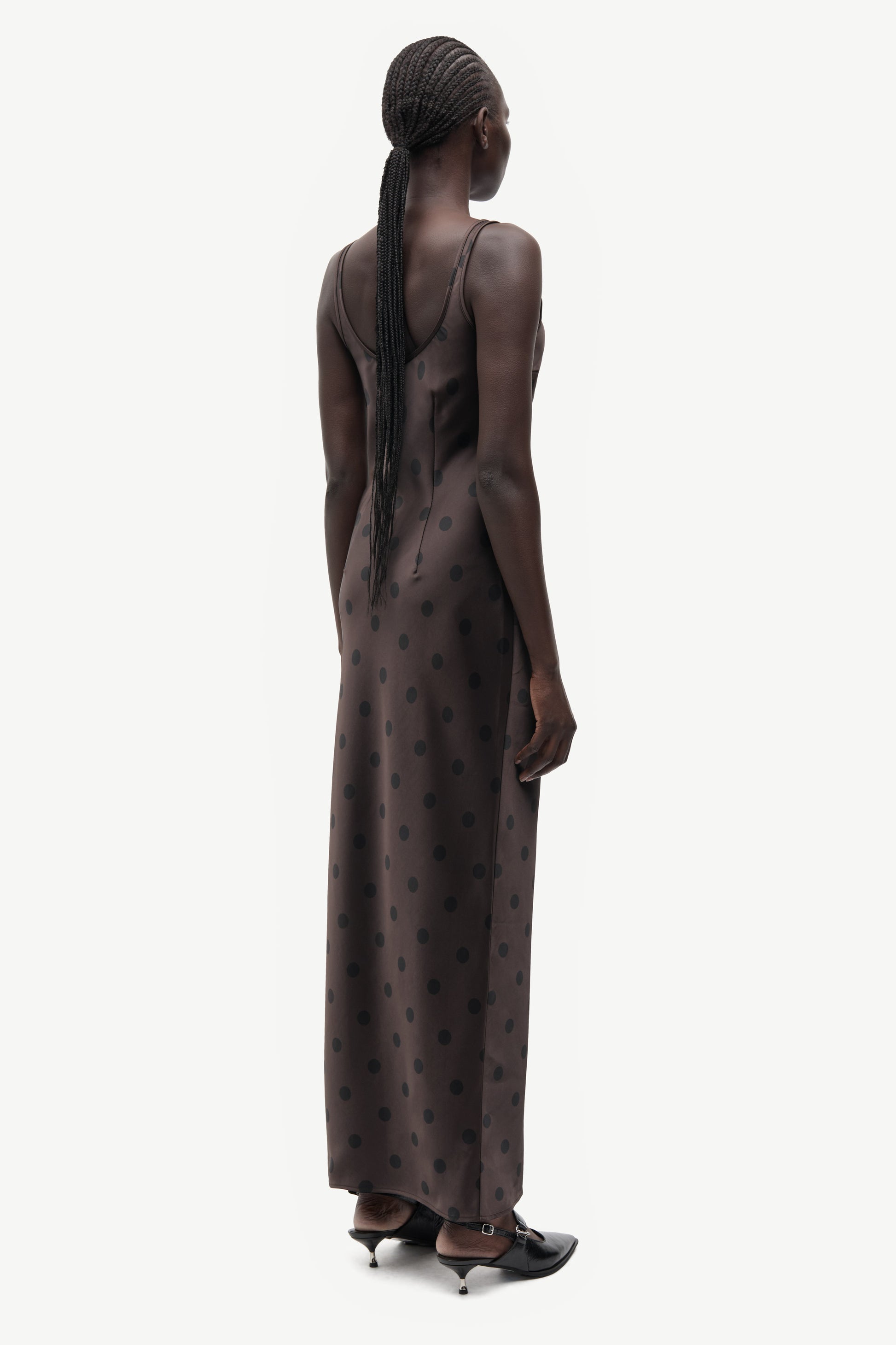 A model shown from the back wearing a dark brown sleeveless maxi dress with a subtle polka dot pattern, paired with black pointed-toe heels, against a plain light gray background.