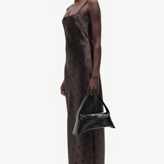 A model is shown in profile wearing a dark brown satin slip dress with a small black polka dot pattern, holding a black handbag and wearing black pointed-toe flats against a plain light gray background.