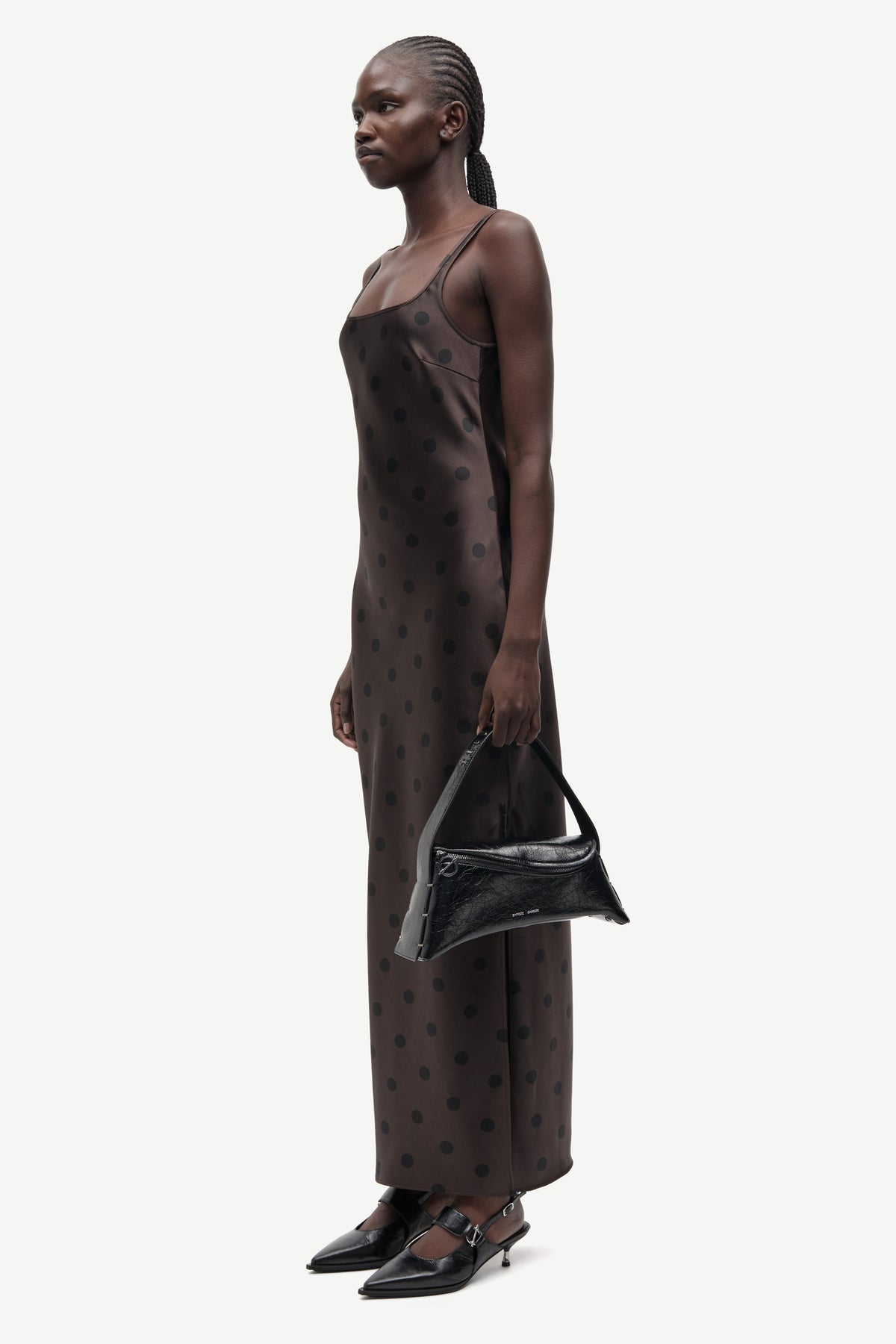 A model is shown in profile wearing a dark brown satin slip dress with a small black polka dot pattern, holding a black handbag and wearing black pointed-toe flats against a plain light gray background.