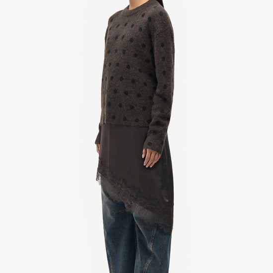A model is shown in profile wearing a dark brown textured knit tunic with a sheer lace overlay and wide-leg blue jeans, set against a plain light gray background.