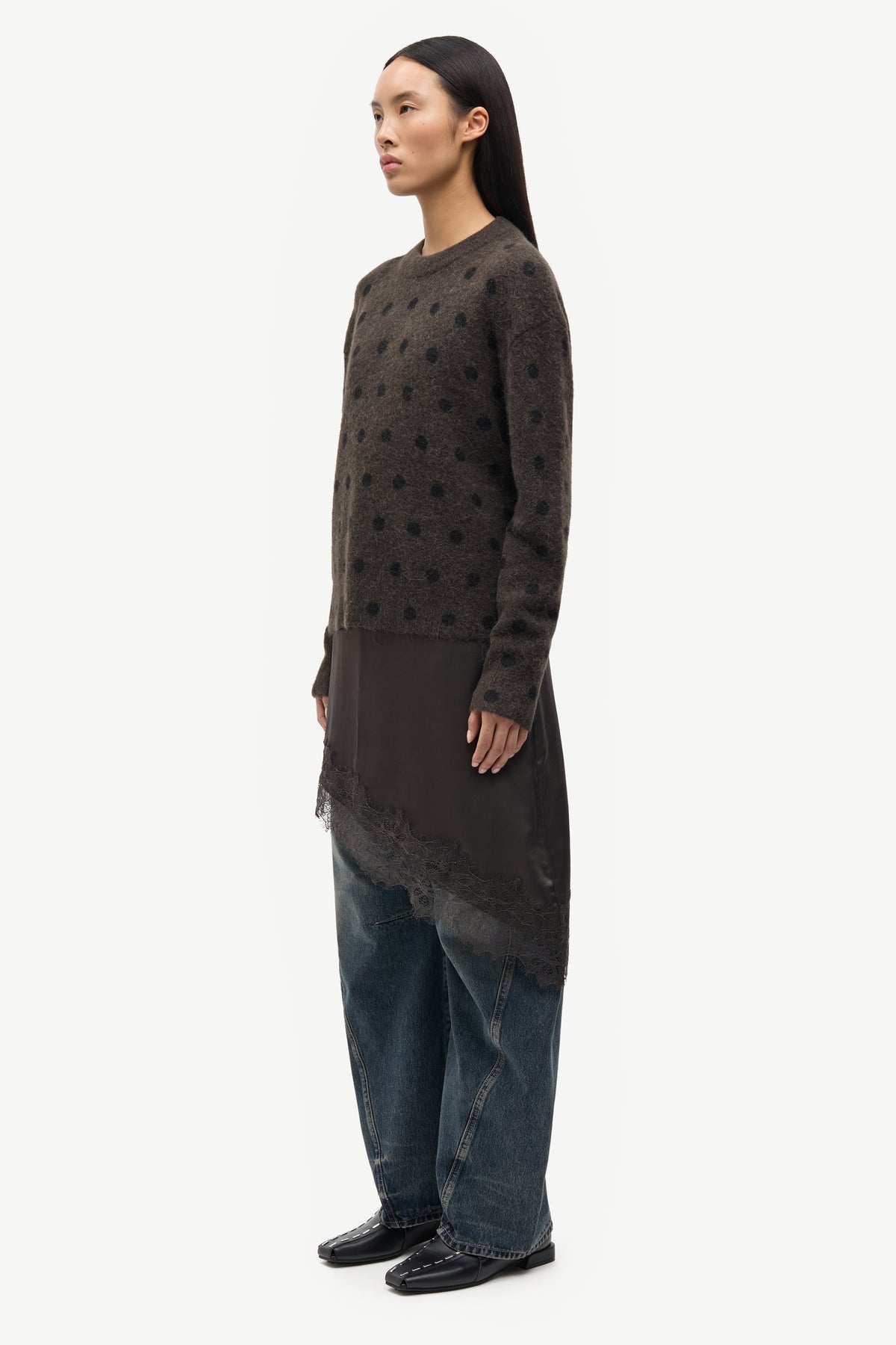 A model is shown in profile wearing a dark brown textured knit tunic with a sheer lace overlay and wide-leg blue jeans, set against a plain light gray background.