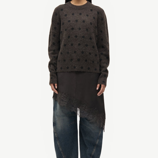 A model stands against a plain white studio background wearing a dark brown long-sleeve knit sweater with a textured dotted pattern, a black asymmetrical draped tunic, wide-leg dark blue jeans, and black shoes.