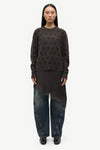 A model stands against a plain white studio background wearing a dark brown long-sleeve knit sweater with a textured dotted pattern, a black asymmetrical draped tunic, wide-leg dark blue jeans, and black shoes.