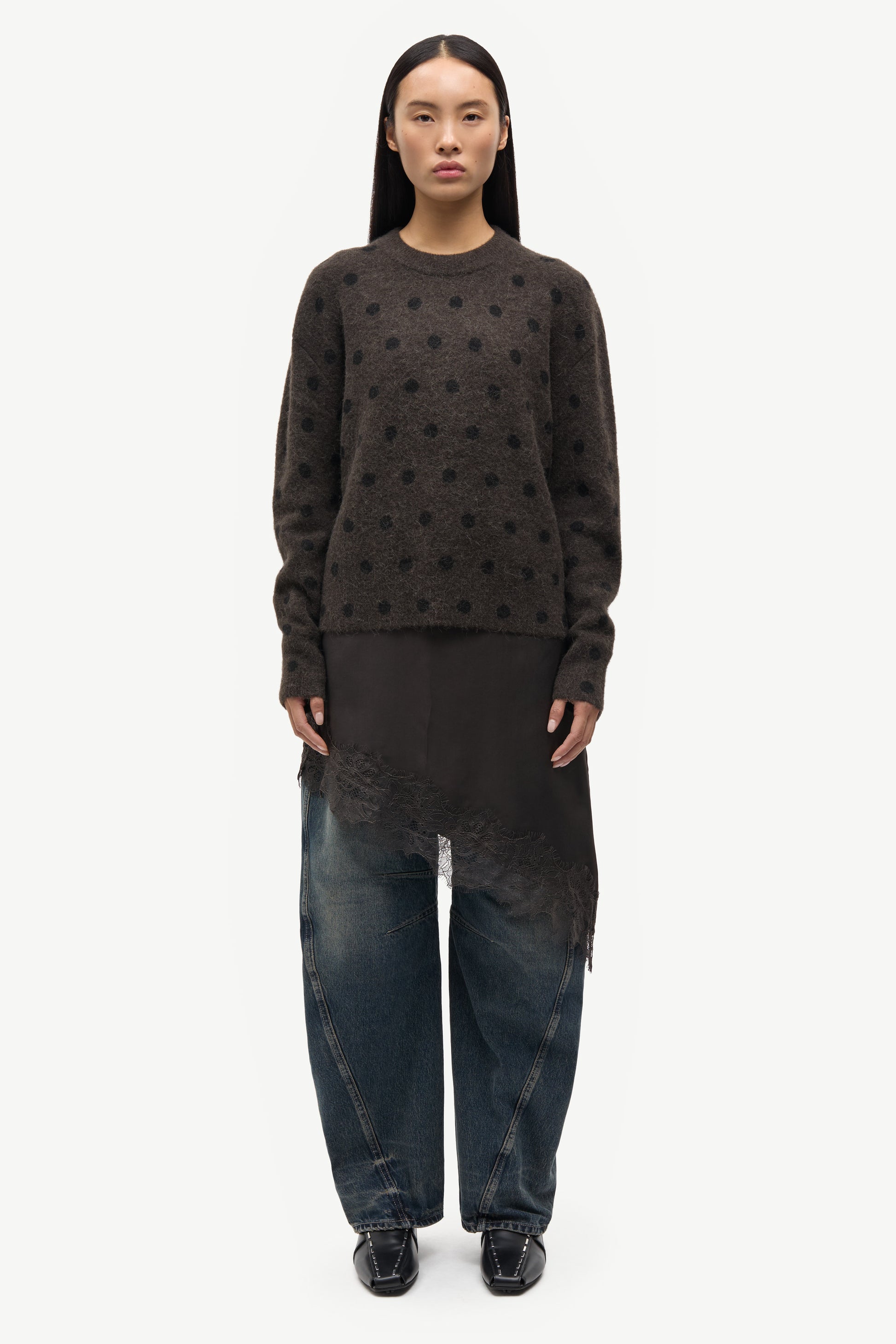 A model stands against a plain white studio background wearing a dark brown long-sleeve knit sweater with a textured dotted pattern, a black asymmetrical draped tunic, wide-leg dark blue jeans, and black shoes.