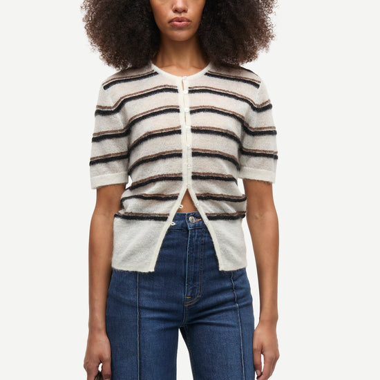 A model with dark curly hair wearing a short-sleeve knit cardigan with horizontal beige, white, and dark brown stripes, paired with blue jeans, against a plain light gray background.