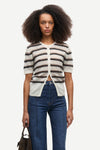 A model with dark curly hair wearing a short-sleeve knit cardigan with horizontal beige, white, and dark brown stripes, paired with blue jeans, against a plain light gray background.