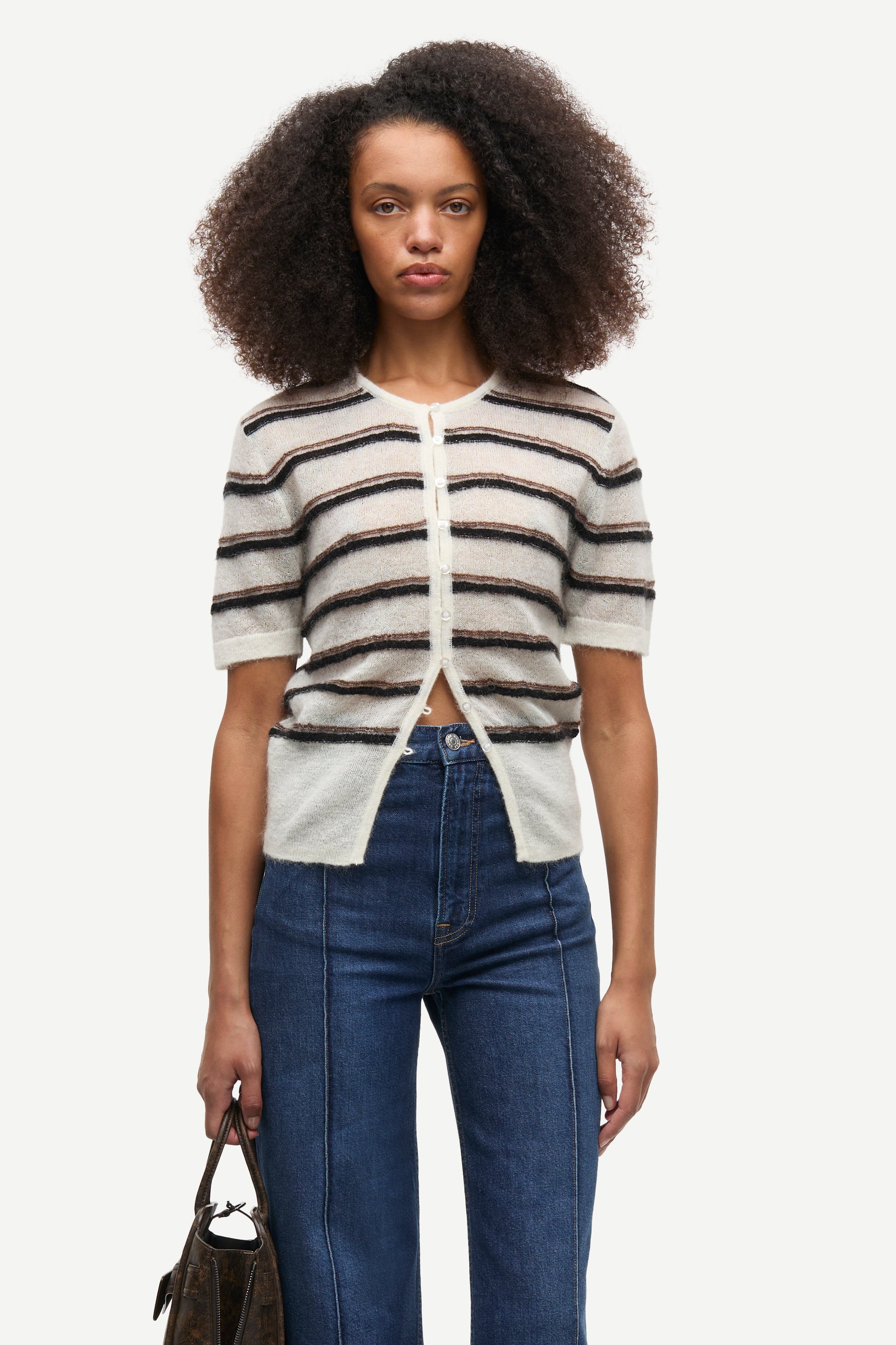 A model with dark curly hair wearing a short-sleeve knit cardigan with horizontal beige, white, and dark brown stripes, paired with blue jeans, against a plain light gray background.