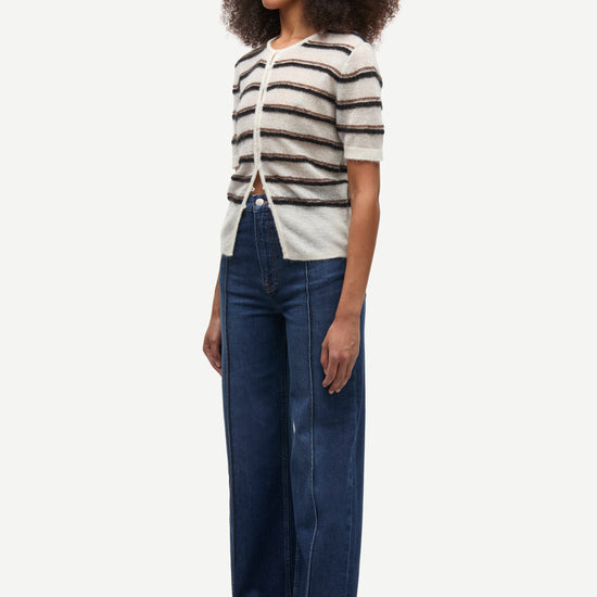 A woman modeling a short-sleeve knit top with horizontal black and off-white stripes, paired with dark blue wide-leg jeans and brown shoes, against a plain light gray background.