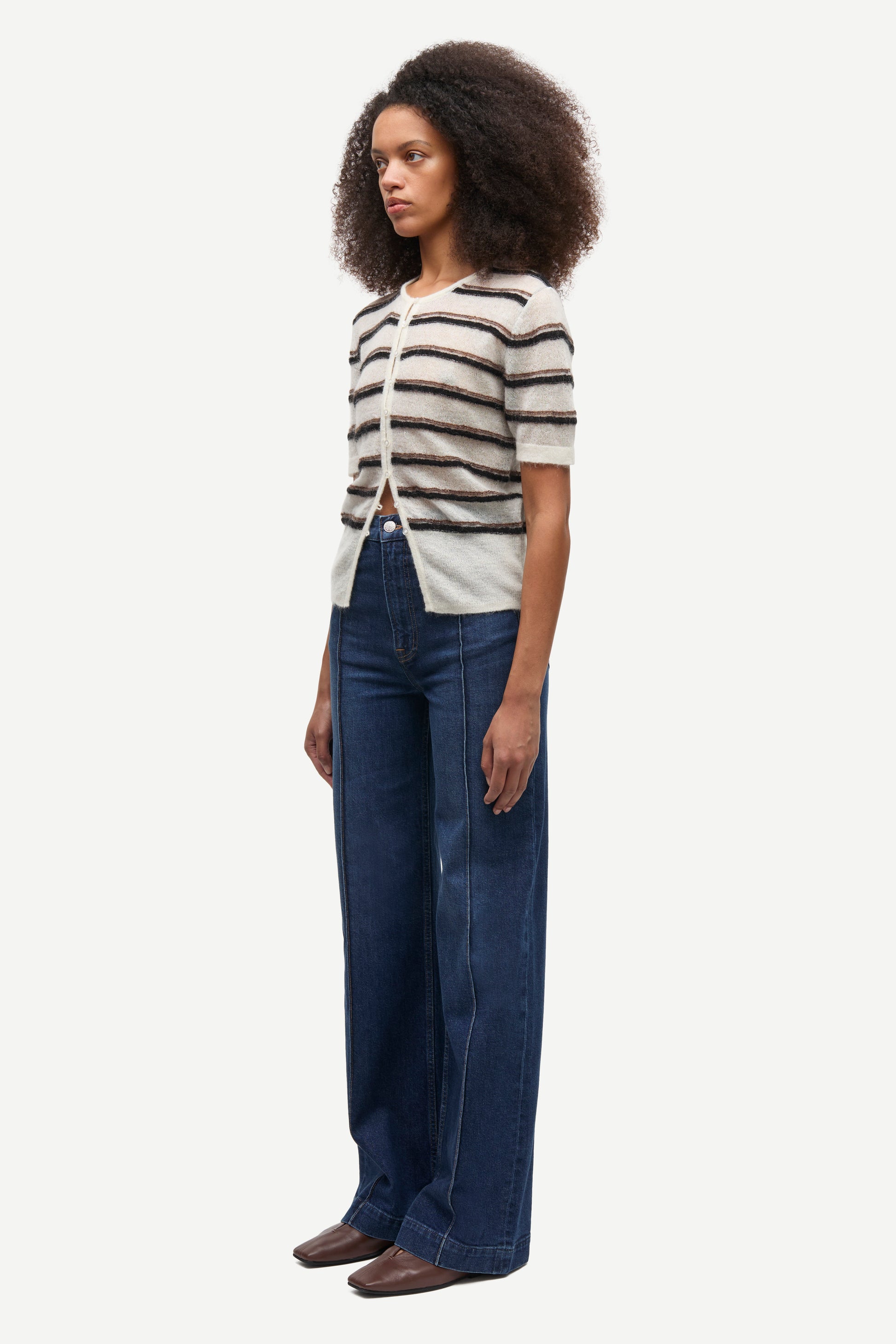 A woman modeling a short-sleeve knit top with horizontal black and off-white stripes, paired with dark blue wide-leg jeans and brown shoes, against a plain light gray background.