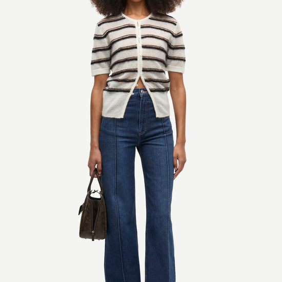 A model stands against a light gray studio background wearing a cream knit short-sleeve cardigan with dark horizontal stripes, blue high-waisted wide-leg jeans, and brown shoes, while holding a dark brown handbag.