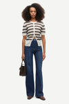 A model stands against a light gray studio background wearing a cream knit short-sleeve cardigan with dark horizontal stripes, blue high-waisted wide-leg jeans, and brown shoes, while holding a dark brown handbag.