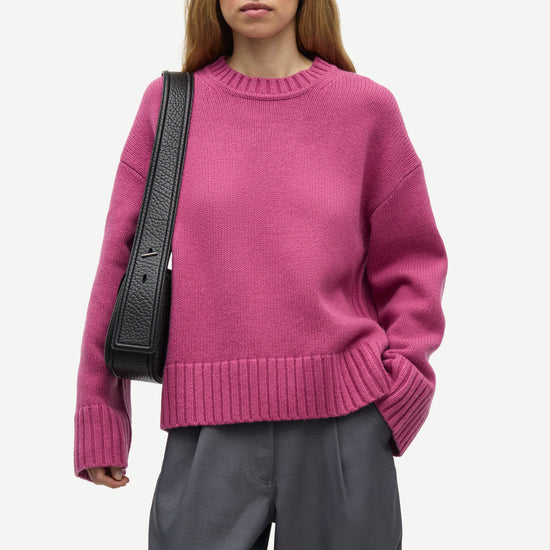 A woman wearing a magenta crewneck knit sweater and dark gray trousers, carrying a black shoulder bag, photographed against a light gray studio background.