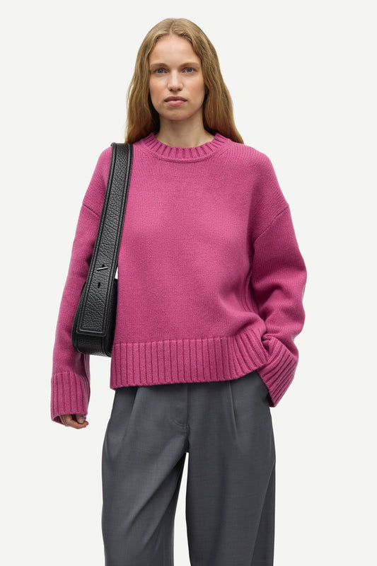 A woman wearing a magenta crewneck knit sweater and dark gray trousers, carrying a black shoulder bag, photographed against a light gray studio background.