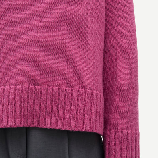Close-up of a person wearing a ribbed knit sweater in a muted magenta-purple color, paired with dark gray trousers, against a plain light background.