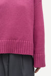 Close-up of a person wearing a ribbed knit sweater in a muted magenta-purple color, paired with dark gray trousers, against a plain light background.