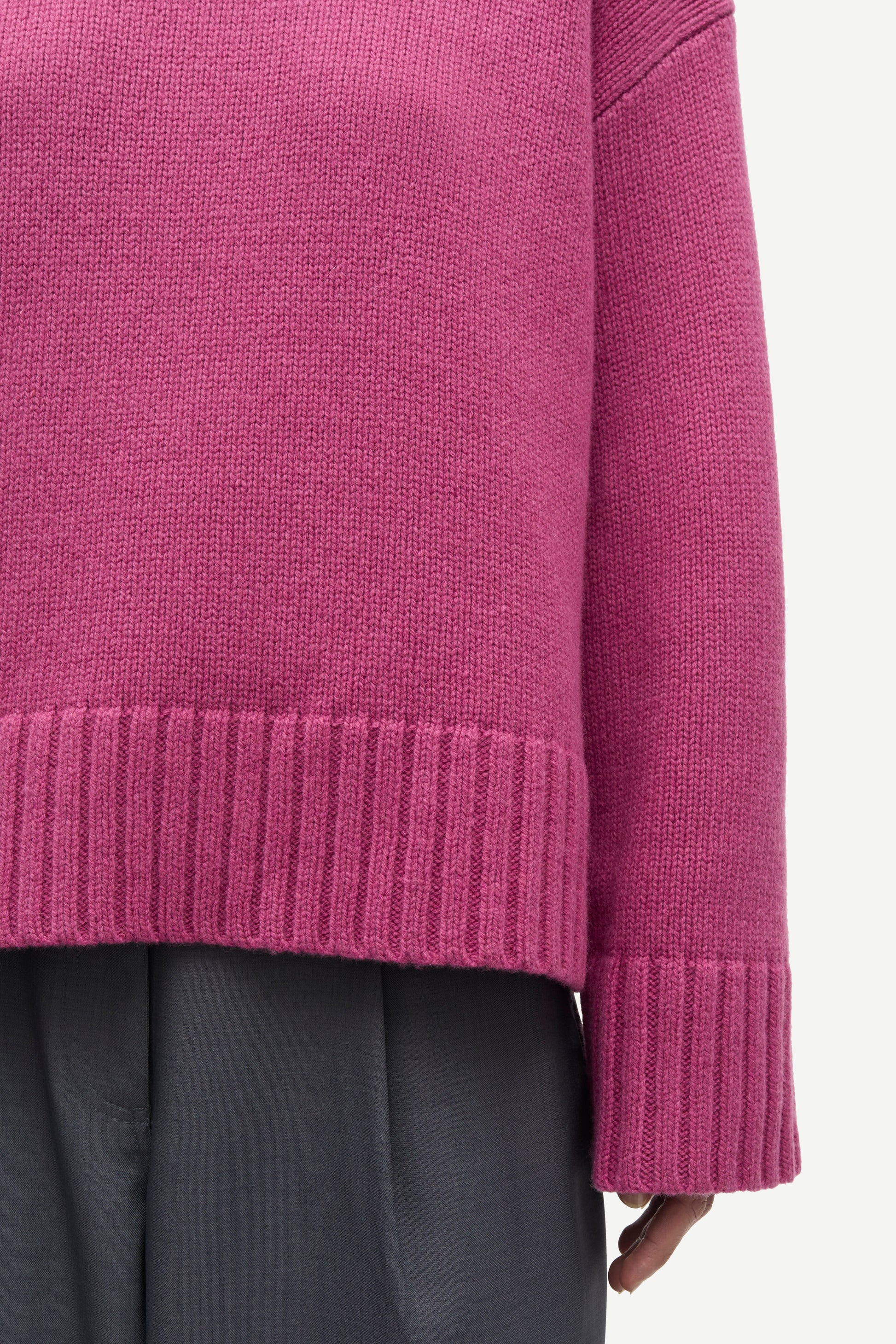 Close-up of a person wearing a ribbed knit sweater in a muted magenta-purple color, paired with dark gray trousers, against a plain light background.