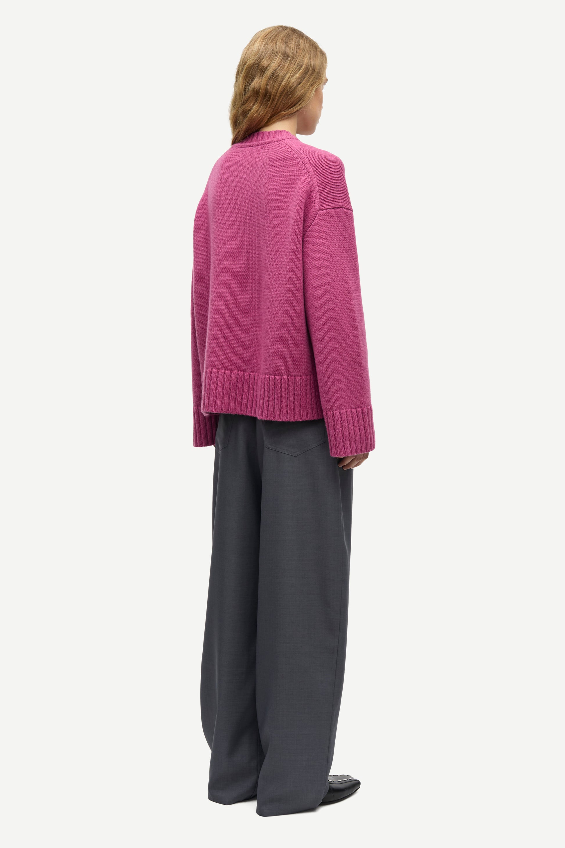 A model shown from the back wearing a magenta knit cardigan and dark gray wide-leg trousers, photographed against a solid off-white background.