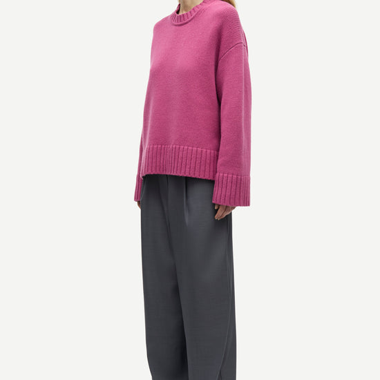 A woman modeling a pink knit sweater and dark gray wide-leg trousers against a plain light gray background.