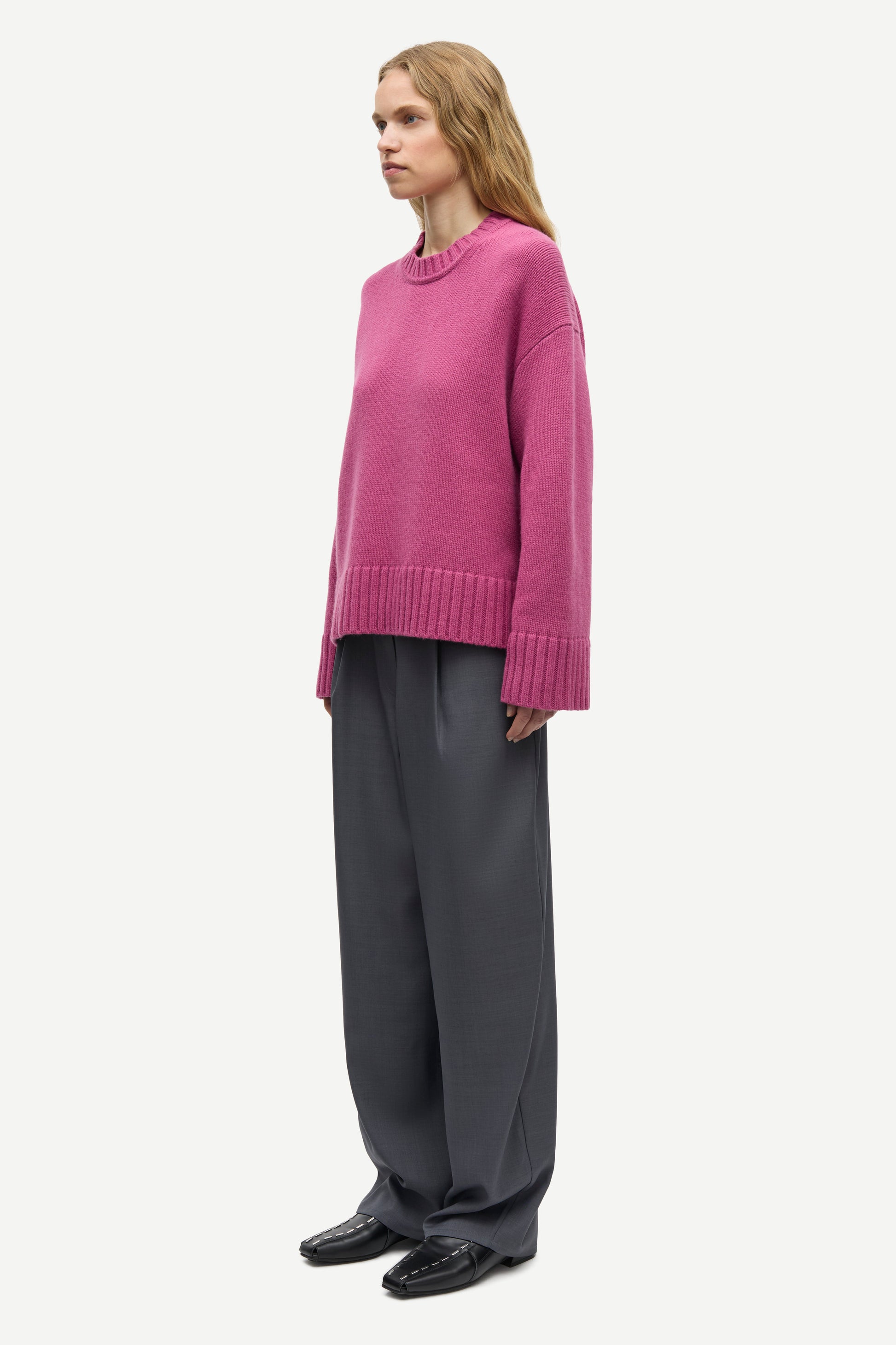 A woman modeling a pink knit sweater and dark gray wide-leg trousers against a plain light gray background.