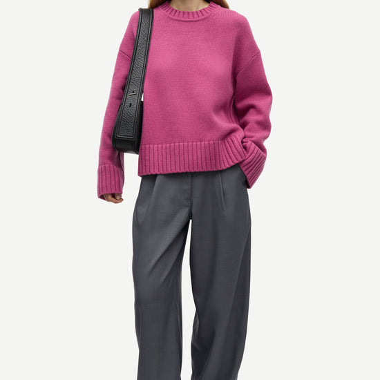 A woman modeling an oversized magenta knit sweater paired with wide-leg charcoal gray trousers and black loafers, carrying a black shoulder bag against a light gray studio background.
