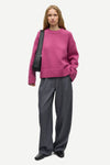 A woman modeling an oversized magenta knit sweater paired with wide-leg charcoal gray trousers and black loafers, carrying a black shoulder bag against a light gray studio background.