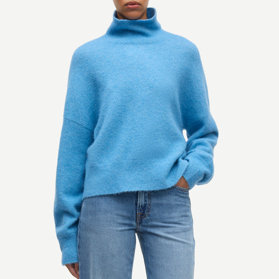 A model wearing a light blue fuzzy turtleneck sweater and light blue jeans, standing against a plain off-white background.
