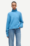 A model wearing a light blue fuzzy turtleneck sweater and light blue jeans, standing against a plain off-white background.