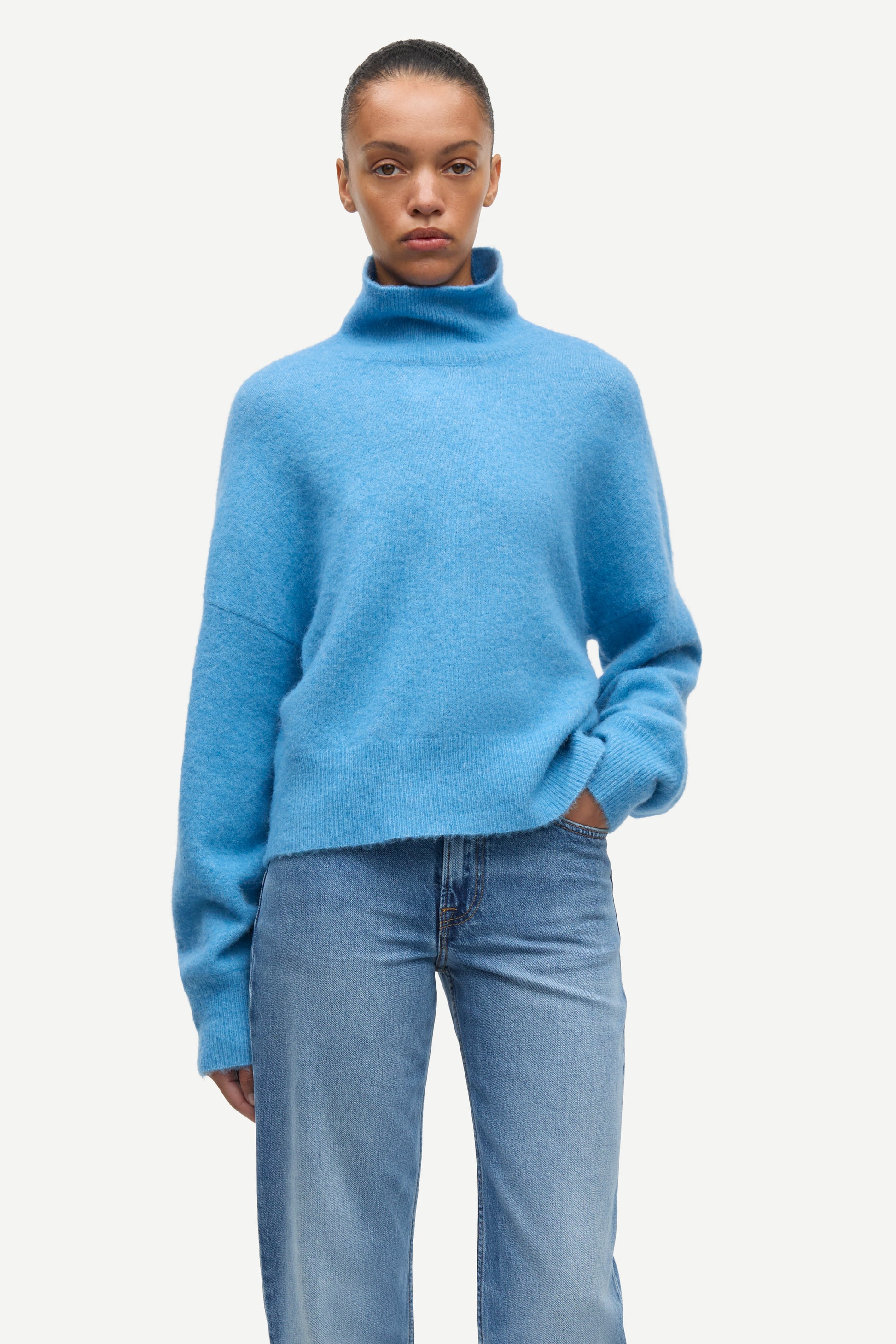 A model wearing a light blue fuzzy turtleneck sweater and light blue jeans, standing against a plain off-white background.