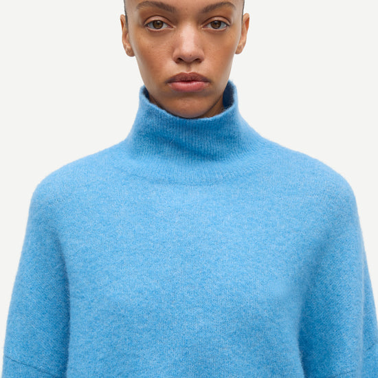 A model wearing a light blue knit turtleneck sweater against a plain off-white background.