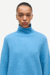 A model wearing a light blue knit turtleneck sweater against a plain off-white background.