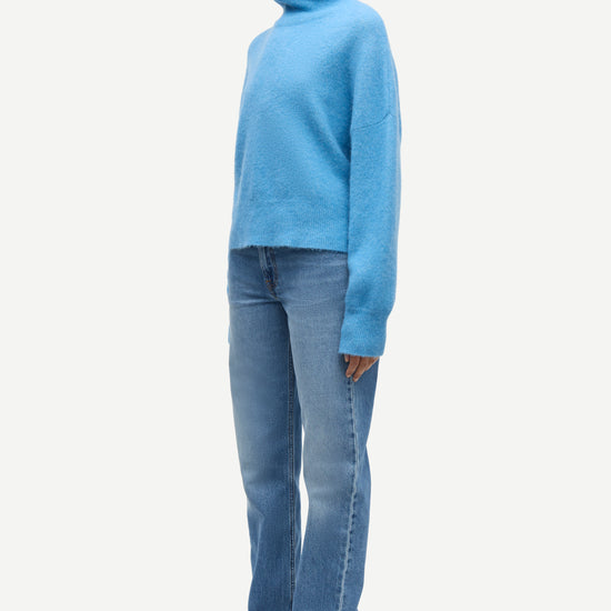 A model wearing a light blue turtleneck sweater, blue jeans, and black leather shoes, shown in profile against a plain light gray background.