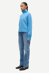 A model wearing a light blue turtleneck sweater, blue jeans, and black leather shoes, shown in profile against a plain light gray background.