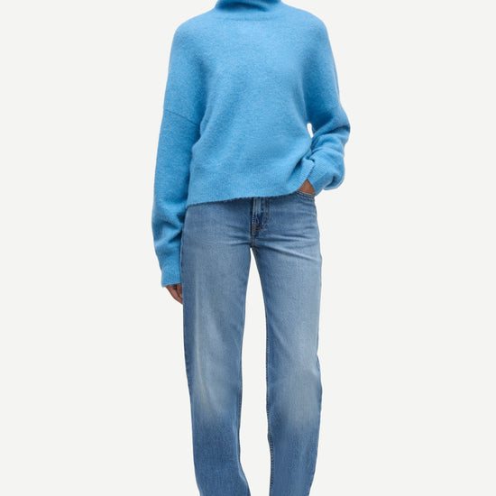 A model wearing a light blue turtleneck sweater and light blue jeans with black shoes, standing against a plain light gray background.