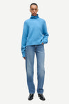A model wearing a light blue turtleneck sweater and light blue jeans with black shoes, standing against a plain light gray background.