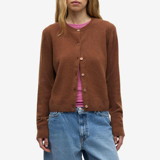 A woman modeling a brown button-up cardigan sweater over a pink top and blue jeans, photographed against a plain light gray studio background.