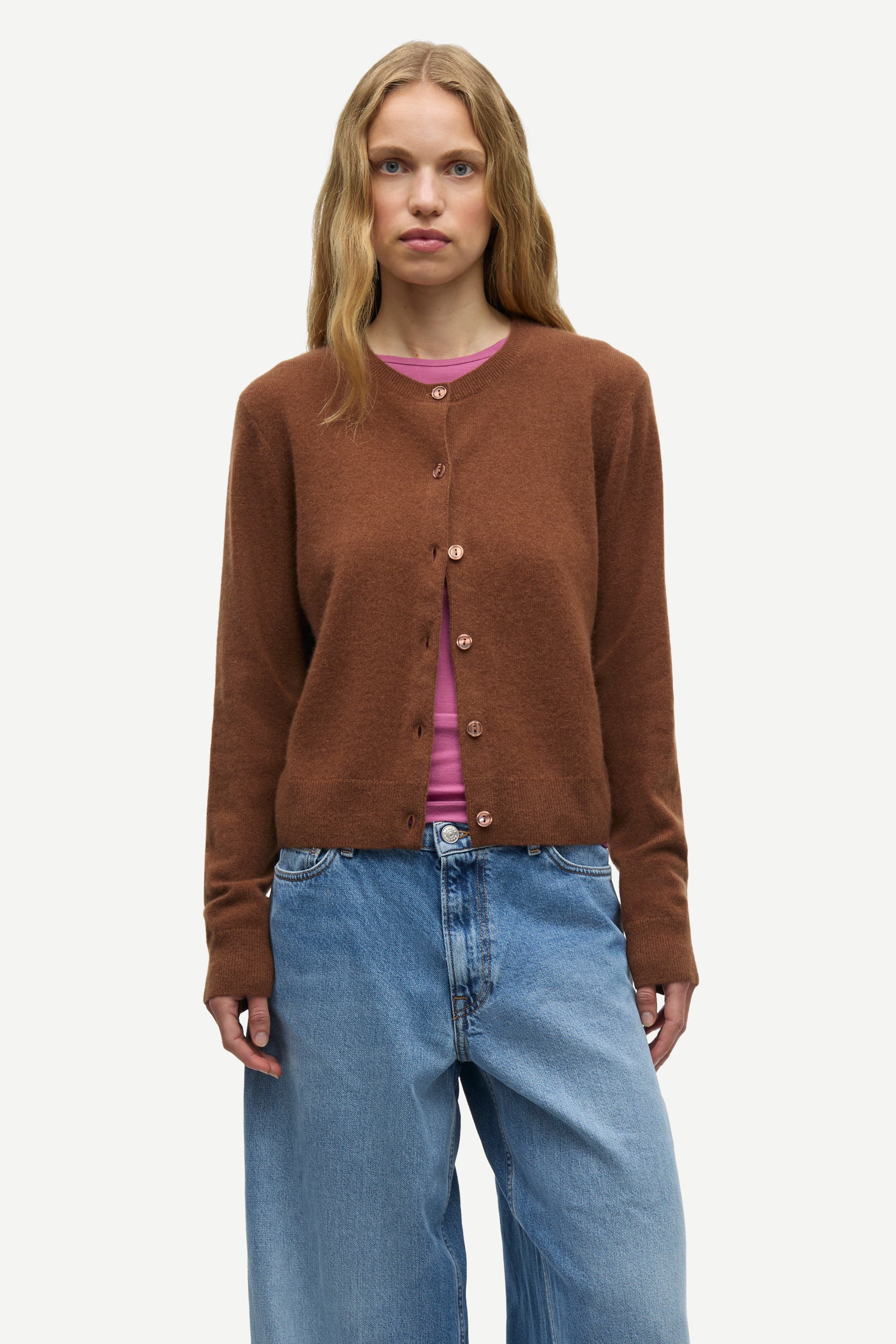 A woman modeling a brown button-up cardigan sweater over a pink top and blue jeans, photographed against a plain light gray studio background.