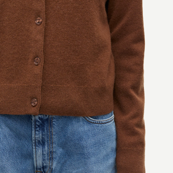 Close-up of a person wearing a brown button-front cardigan sweater with blue jeans against a plain light background.
