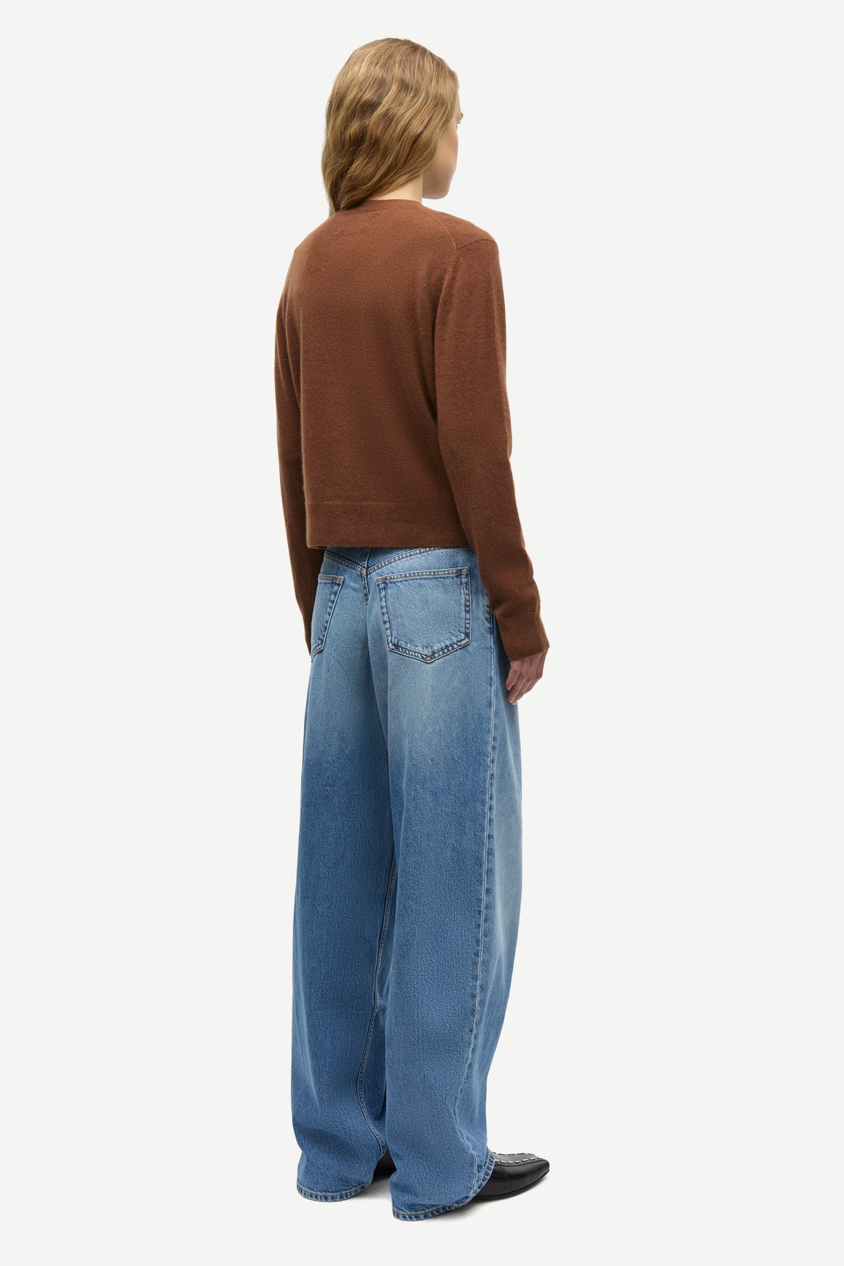 A model shown from the back wearing a brown cropped long-sleeve knit top and wide-leg light blue denim jeans, photographed against a solid off-white background.