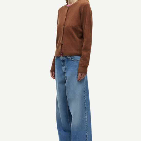 A woman modeling a brown long-sleeve knit cardigan and wide-leg blue jeans against a plain light gray studio background.