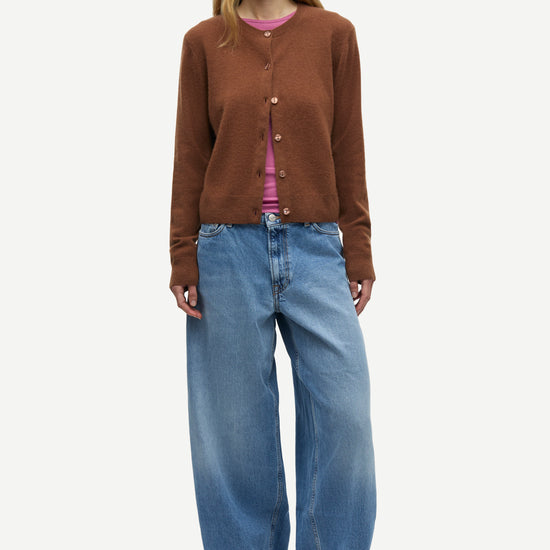 A model stands against a light gray studio background wearing a brown button-front cardigan over a pink top, paired with wide-leg blue jeans and black shoes.