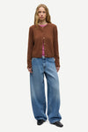A model stands against a light gray studio background wearing a brown button-front cardigan over a pink top, paired with wide-leg blue jeans and black shoes.