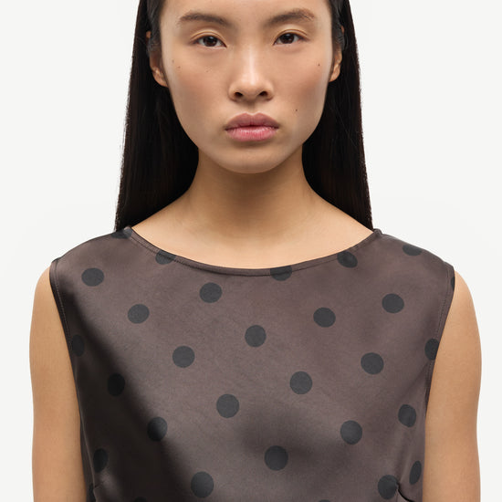 A woman wearing a sleeveless dark brown top with black polka dots, shown from the chest up against a plain light background.
