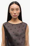 A woman wearing a sleeveless dark brown top with black polka dots, shown from the chest up against a plain light background.