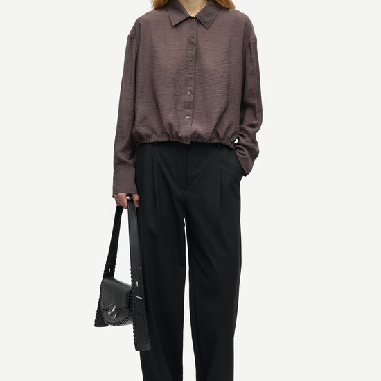 A model stands against a light gray studio background wearing a dark brown textured long-sleeve button-up shirt, black wide-leg trousers, and black shoes, while holding a black shoulder bag.